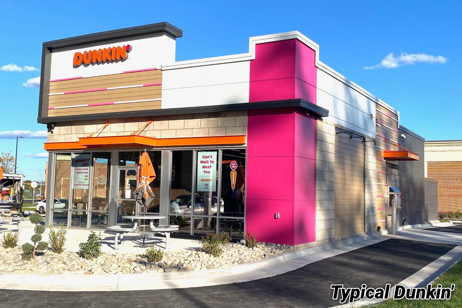 Exterior photo of a typical Dunkin' restaurant — a pink, orange, and tan building with the Dunkin' logo and a small patio.